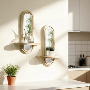 May include: Two wooden wall mirrors with shelves, each featuring an arched mirror design. One shelf holds a glass vase with greenery, the other holds potted plants. The mirrors are mounted on a white wall, with a terracotta pot of greenery in the foreground.