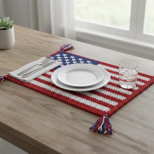 May include: A crocheted placemat in the style of the American flag, featuring red and white stripes and a blue corner with white stars. Two white plates, silverware, and a glass of water are on the placemat. Tassels adorn each end.