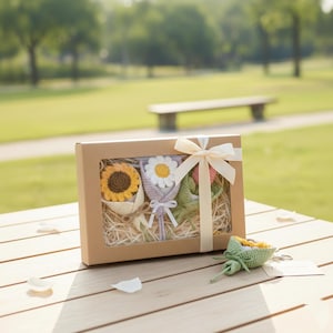 May include: A gift box with three crocheted flower bouquets: a sunflower, a daisy, and a pink flower. The box is tied with a cream-colored ribbon. A small crocheted sunflower keychain is also visible.