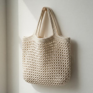 Peut inclure: Un sac fourre-tout en crochet crème à tissage ouvert, avec deux poignées robustes. Le sac est suspendu à une cheville en bois contre un mur blanc. Le design du sac présente un motif en forme de grille, et sa couleur neutre le rend polyvalent.