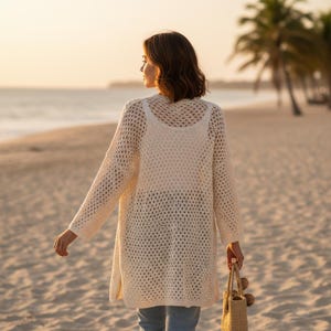 May include: A woman on a sandy beach wearing a cream-coloured, open-weave cardigan over a matching vest top. The cardigan has long sleeves and a knee-length hem. She carries a woven tote bag. Palm trees and the sea are in the background.