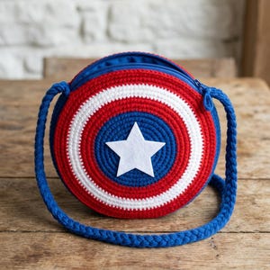 May include: A round, crocheted bag in red, white, and blue, resembling a shield. The bag features a white star in the center and a blue zipper. It has a blue strap for carrying. The bag is sitting on a wooden surface.