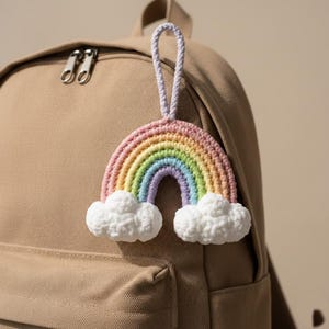 Puede incluir: Un llavero de arcoíris tejido a ganchillo con colores pastel de rosa, amarillo, verde, azul y morado. El arcoíris tiene nubes blancas y esponjosas en la parte inferior y un lazo lavanda para colgar. El llavero está sujeto a una mochila beige.