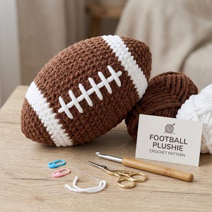 Pode incluir: Um boneco de futebol americano de crochê marrom e branco com costuras brancas, ao lado de fios marrons e brancos, uma agulha de crochê, tesouras e um cartão que diz "FOOTBALL PLUSHIE CROCHET PATTERN".