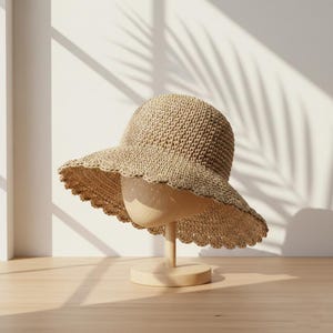 Puede incluir: Un sombrero de sol beige tejido con un borde festoneado, exhibido en un soporte de madera. El sombrero tiene una copa redondeada y un ala ancha. El soporte del sombrero es de madera clara y tiene una base circular. El fondo presenta sombras de una planta.