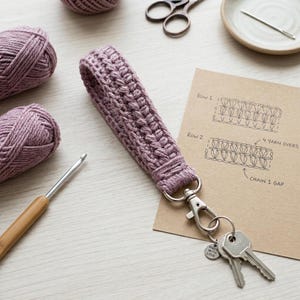 May include: A handmade, mauve-colored crochet keychain wrist strap with a silver clasp and attached keys. The strap is next to yarn, a crochet hook, scissors, and a diagram. The tag on the keychain says "MADE WITH LOVE."