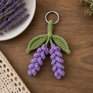 Puede incluir: Un llavero de ganchillo con dos tallos de flores de lavanda moradas con hojas verdes unidos a una anilla de metal plateado. Las flores de lavanda tienen una textura con patrón de nudos y dos hojas verdes situadas sobre las flores.