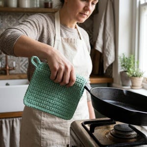 Op de afbeelding: Een lichtgroene, vierkante gehaakte pannenlap met een ophanglus. De pannenlap wordt gebruikt om een zwarte gietijzeren koekenpan vast te houden. De keuken omvat een fornuis, een gootsteen en een raam.