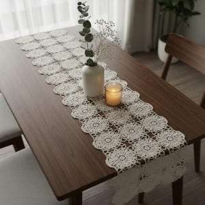 Może przedstawiać: Kremowy, szydełkowy bieżnik na stół z kwiatowym wzorem, umieszczony na ciemnym drewnianym stole. Na stole znajduje się również biały wazon z zielenią i zapalona świeca w szklanym świeczniku. Bieżnik dodaje elegancji.