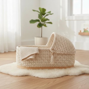 May include: A cream-colored, crocheted baby bassinet with a hood and a small blanket. The bassinet sits on a wooden base and is decorated with a tassel. The bassinet is placed on a white, fluffy rug.