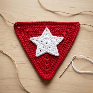 May include: A handmade red crochet banner with a white star applique. The banner is triangular and has a textured surface. A needle and white thread are visible, suggesting a DIY craft project.