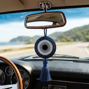 May include: A blue and white crochet evil eye car charm hanging from a car's rearview mirror. The charm has a blue tassel and is set against a scenic background of a road and mountains.