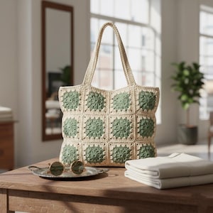 Puede incluir: Un bolso tote de crochet beige y verde con un patrón cuadrado. El bolso tiene una forma estructurada con un asa. Unas gafas de sol redondas descansan sobre una pequeña bandeja plateada. Una pila de tela blanca doblada está al lado del bolso.
