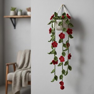 May include: A handmade, off-white crochet hanging planter with red flowers and green leaves. The planter is suspended by a white rope and hangs against a light gray wall. The cascading flowers and leaves add a touch of nature to the decor.