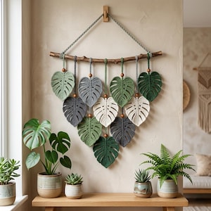 Könnte beinhalten: Handgefertigter Wandbehang mit gehäkelten Monstera-Blättern in Grün-, Grau- und Cremetönen. Die Blätter sind an einem natürlichen Ast mit Holzperlen aufgehängt. Die Wanddekoration ist an einer hellen Wand befestigt und schafft eine natürliche, böhmische Ästhetik.