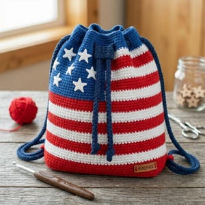May include: A handmade crochet drawstring bag with a patriotic design. The bag features a blue top with white stars and red and white stripes, resembling the American flag. It has blue drawstrings and a small "HANDMADE" tag.