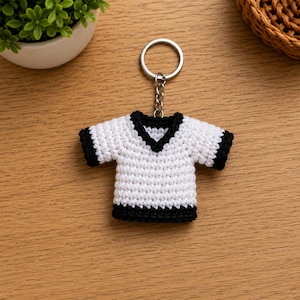 May include: A crochet keyring shaped like a small t-shirt. The shirt is primarily white with a black v-neck collar, black sleeve cuffs, and a black hem. It is attached to a silver-coloured metal split ring by a short silver-coloured metal chain.