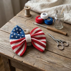 May include: A handmade crochet bow tie in red, white, and blue, resembling the American flag. The bow tie features white stars on a blue field and red and white stripes. Crafting supplies, including yarn, scissors, and a crochet hook, are arranged around the bow tie.