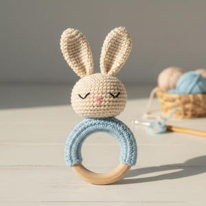 Puede incluir: Un sonajero de conejo de ganchillo hecho a mano. El conejo tiene una cabeza beige claro, orejas largas y ojos cerrados. Se asienta sobre un anillo de ganchillo azul claro sujeto a un anillo de madera. El fondo incluye hilo y una cesta.
