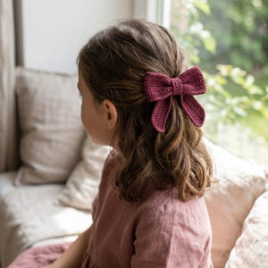 May include: A dark magenta crochet hair bow featuring a textured, hand-stitched yarn construction with a central knot detail and two downward-pointing loops, secured to the hair to hold a section of wavy brown hair in place.