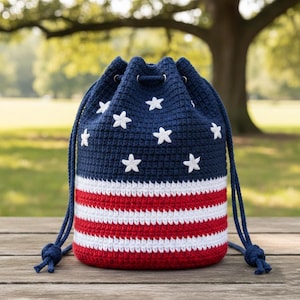 May include: A navy blue and red crochet drawstring bag with white star and stripe details. The bag features a navy blue drawstring closure and is designed with the American flag in mind. The bag is sitting on a wooden surface.