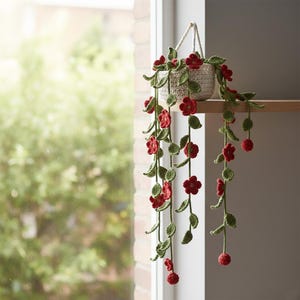 May include: A crocheted hanging basket with trailing vines and red flowers. The beige basket hangs from a white cord. The vines feature green leaves and red flowers and buds. The plant is displayed on a wooden shelf.