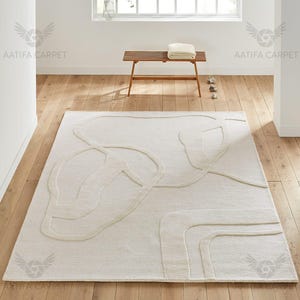 May include: A cream-coloured rectangular rug with an abstract pattern of raised lines. The rug is laid on a wooden floor, and the design features curved shapes. The room is bright, with a bench and window visible in the background.