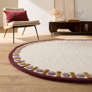 May include: A round, off-white rug with a burgundy border. The border features a repeating pattern of gold and purple shapes. The rug is placed on a hardwood floor, with a chair and side table in the background.