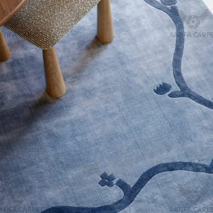 May include: A blue area rug with a floral branch design in a darker shade of blue. The rug is placed under a wooden table and chair. The chair has wooden legs and a patterned seat. The rug's design is a stylized floral pattern.