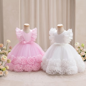 Może przedstawiać: Dwie formalne sukienki, jedna jasnoróżowa, druga biała. Obie mają dekolt w serek, falbaniaste rękawy i kokardę w talii. Spódnice są z tiulu z dużymi, różokształtnymi zdobieniami na dole.