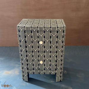 May include: A rectangular bedside table featuring two drawers with a black and white geometric pattern. The surface is covered in a repeating arched line design. Each drawer is equipped with a small, round, off-white knob for opening.