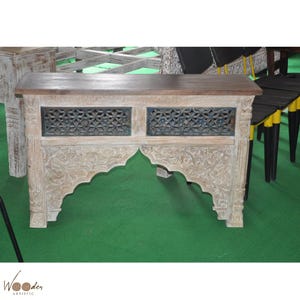 以下が含まれることがあります： 濃い茶色の天板と、精巧に彫刻されたオフホワイトのベースが特徴の木製コンソールテーブル。ベースには花の模様と、幾何学模様の装飾パネルが2つあります。テーブルの幅は約122cmです。