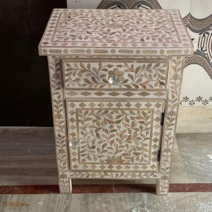 May include: A rectangular bedside table featuring an intricate floral inlay pattern in shades of cream and light brown. The piece includes one top drawer and one lower cabinet door, both equipped with clear, faceted, round glass knobs.