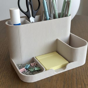 May include: A beige desk tidy with several compartments. It contains pens, scissors, a glue stick, paper clips, and sticky notes. The organiser has a ribbed texture and is on a wooden surface.