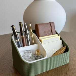 May include: A green and white desk tidy containing pens, a small notebook, stationery, and a gold feather. The organiser has a ribbed exterior and is on a wooden surface. A white vase is in the background.