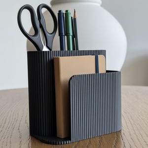 May include: A black desk tidy with vertical ribbed detailing. It contains black scissors, pens, a pencil, and a tan notebook with a blue elastic band. The organiser sits on a wooden surface.