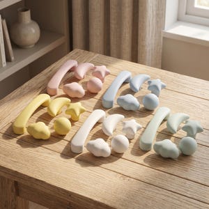 May include: A collection of cabinet hardware sets in pastel colors, including pink, blue, yellow, white, and mint green. Each set features a long curved handle, a crescent moon knob, a star-shaped knob, a cloud-shaped knob, and a spherical knob.
