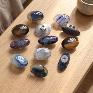 Op de afbeelding: Een collectie van elf ovale kastknoppen gemaakt van gepolijste natuurstenen en mineralen. Elke knop heeft unieke patronen, waaronder gebandeerde agaat, tijgeroog en gebarsten texturen in tinten blauw, bruin en paars.