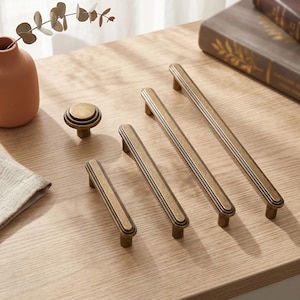May include: A collection of antique brass hardware including one round cabinet knob and three elongated drawer pulls of varying lengths. Each piece features a recessed center panel with a raised border and a smooth, weathered metallic finish.