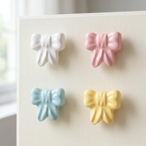 May include: A set of four ceramic cabinet knobs shaped like bows, featuring a glossy finish in white, pastel pink, light blue, and soft yellow. Each knob has a central knot and two hanging loops, providing a decorative hardware option.