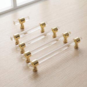 Puede incluir: Un conjunto de herrajes para muebles que consta de un pomo pequeño y cuatro tiradores largos para cajones. Cada pieza presenta una barra cilíndrica de acrílico transparente con soportes metálicos en tono dorado pulido.