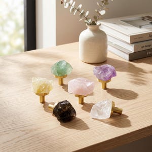 May include: A collection of six cabinet knobs featuring raw, unpolished gemstone tops in yellow, green, light pink, purple, dark brown, and clear white. Each stone is securely mounted onto a cylindrical, brushed gold-tone metal base.