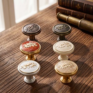 May include: Six round cabinet knobs arranged in two columns. Each knob features a raised leaf motif on the circular face. The finishes include dark bronze, polished gold, silver, cream, and marbled red and beige tones with metallic edges.