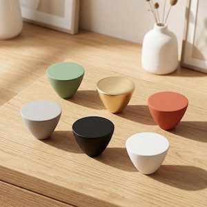 May include: A collection of six cabinet knobs in a conical shape, featuring a smooth finish in various colours including sage green, brushed gold, terracotta orange, light grey, matte black, and crisp white, arranged on a light wood surface.