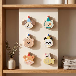 May include: A rectangular off-white board featuring six wooden animal-shaped magnets. The magnets depict a dog, a cat, a bear, a panda, a person, and a fish, each wearing a colorful party hat. The board is displayed vertically on a wooden shelf.