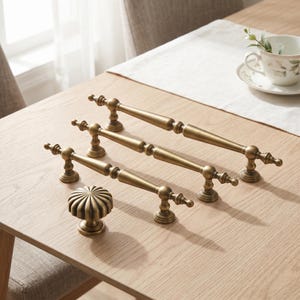 Könnte beinhalten: Ein Set aus antiken Messingbeschlägen, bestehend aus drei länglichen Schrankgriffen mit spitz zulaufendem, spindelartigem Design und einem runden, geriffelten Schrankknopf. Jedes Teil hat eine glatte Metalloberfläche mit dunklen Details.