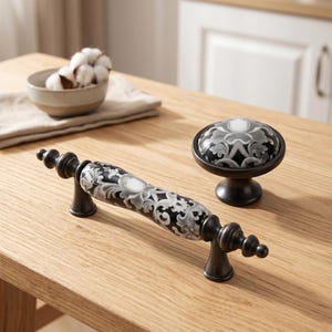 Può includere: Una maniglia per mobili e un pomello rotondo abbinato, entrambi caratterizzati da un motivo floreale bianco e nero su un centro in ceramica con estremità in metallo scuro. La maniglia ha un design lungo e curvo, mentre il pomello è circolare.