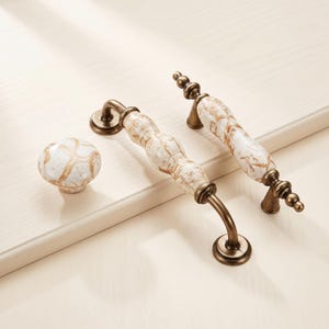May include: A set of cabinet hardware including one round knob and two arched drawer pulls. Each piece features a white ceramic surface with a brown marbled pattern and antique brass-toned metal accents at the base and ends of the handles.