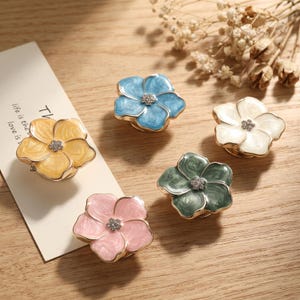 May include: Five flower-shaped decorative pins in yellow, blue, white, green, and pink. Each pin features a swirling enamel finish, gold-toned metal edges, and a small cluster of clear crystals at the center. A white card with text is visible.