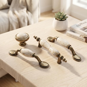May include: A set of furniture hardware including one round knob and two arched drawer pulls. Each piece features a white marble-patterned ceramic center with antique brass-toned metal ends and mounting bases, showcasing a smooth finish.
